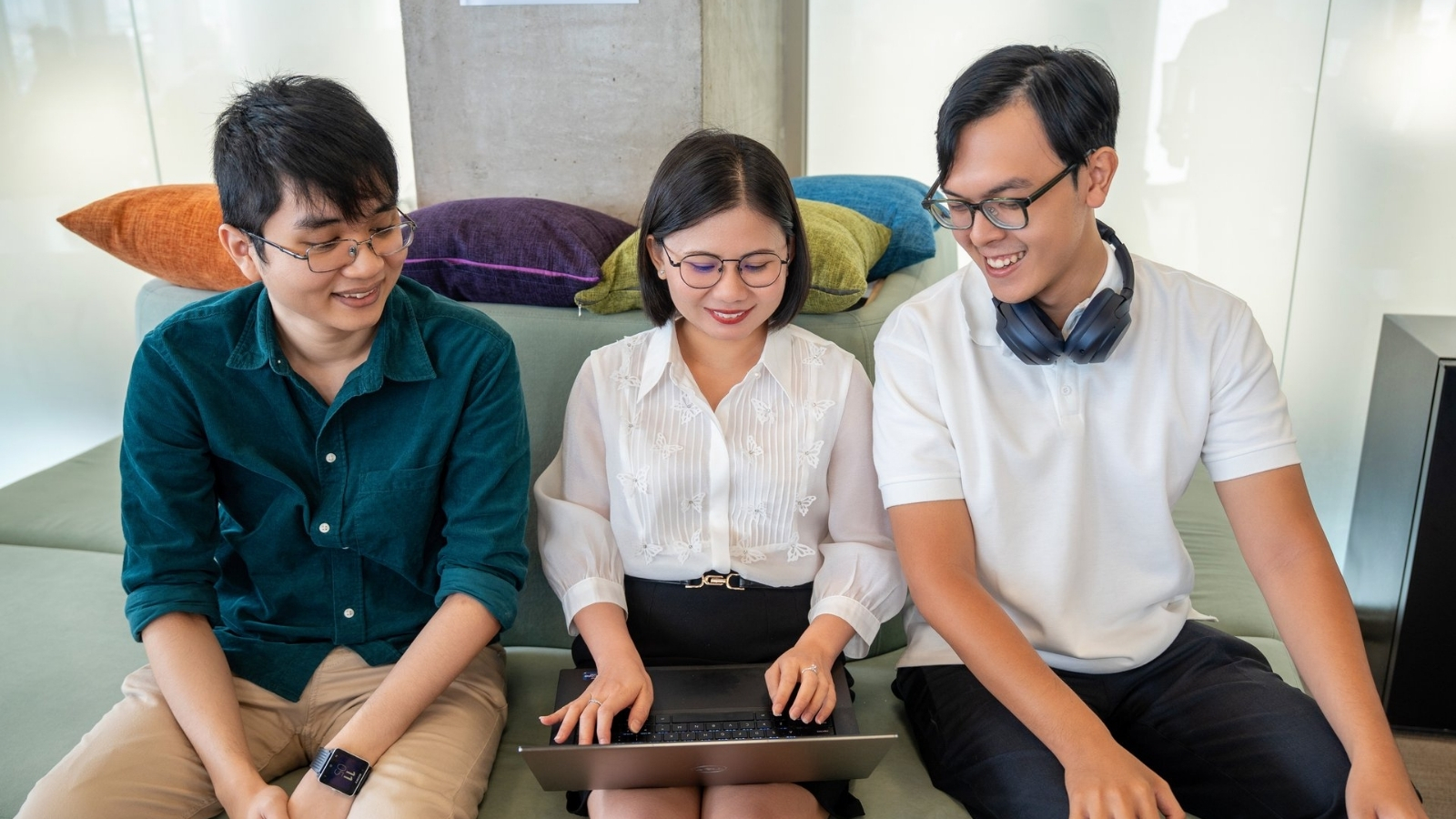 Three colleagues sitting together on a sofa, smiling as they review content on a laptop during a collaborative work session.