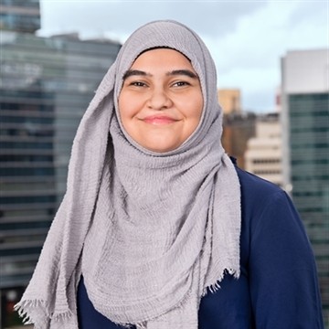Portrait of a smiling woman wearing a light grey hijab and a navy-blue top, standing outdoors with modern office buildings blurred in the background.