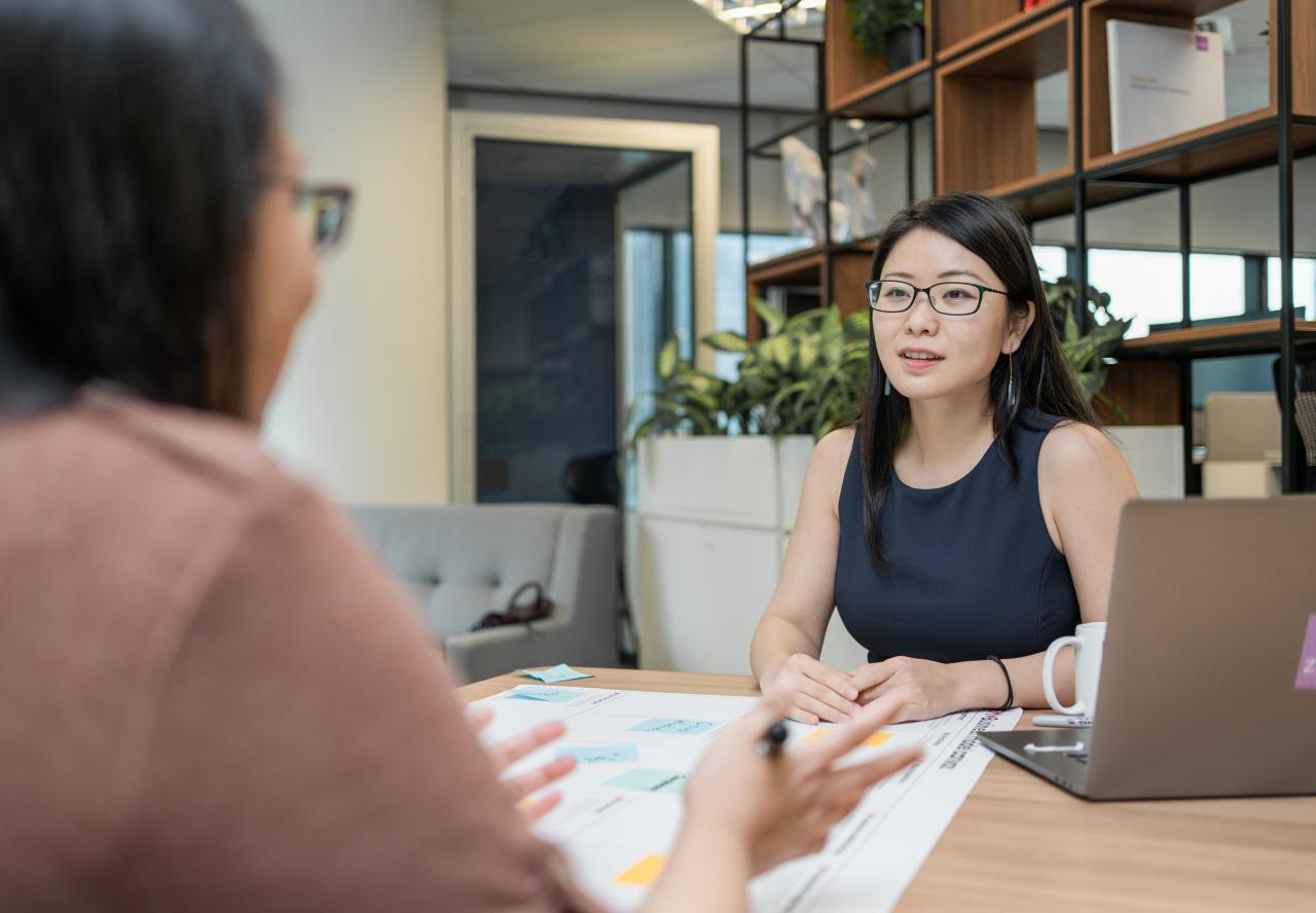 A woman wearing glasses is sitting in a modern office meeting space, engaged in conversation with a colleague across the table.