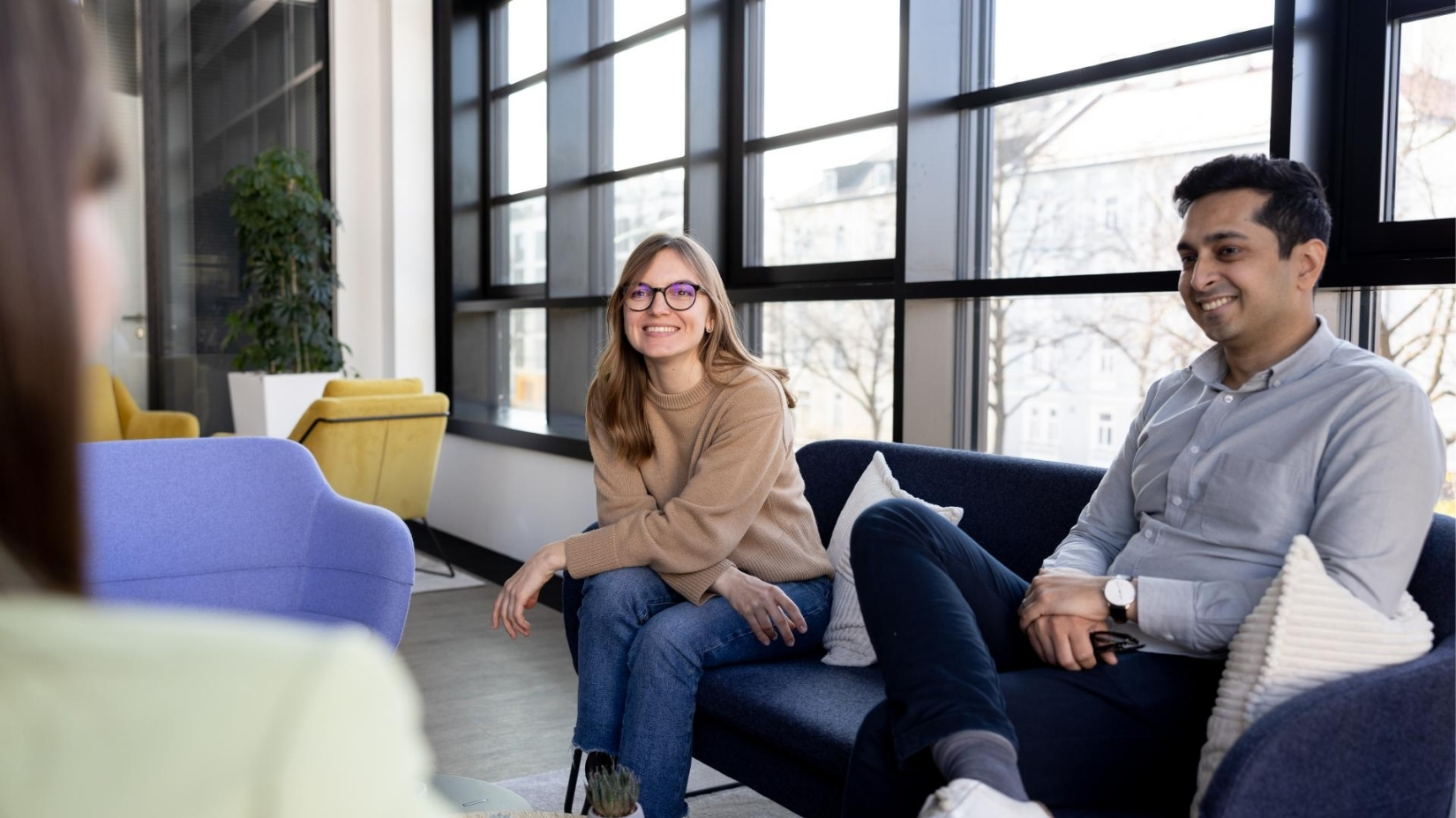 Two colleagues sitting on a sofa, smiling as they chat with another colleague in a bright office lounge.