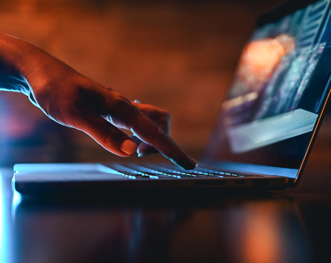 Close-up of a person’s hand typing on a laptop keyboard, illuminated by blue and orange lighting, with code visible on the screen.