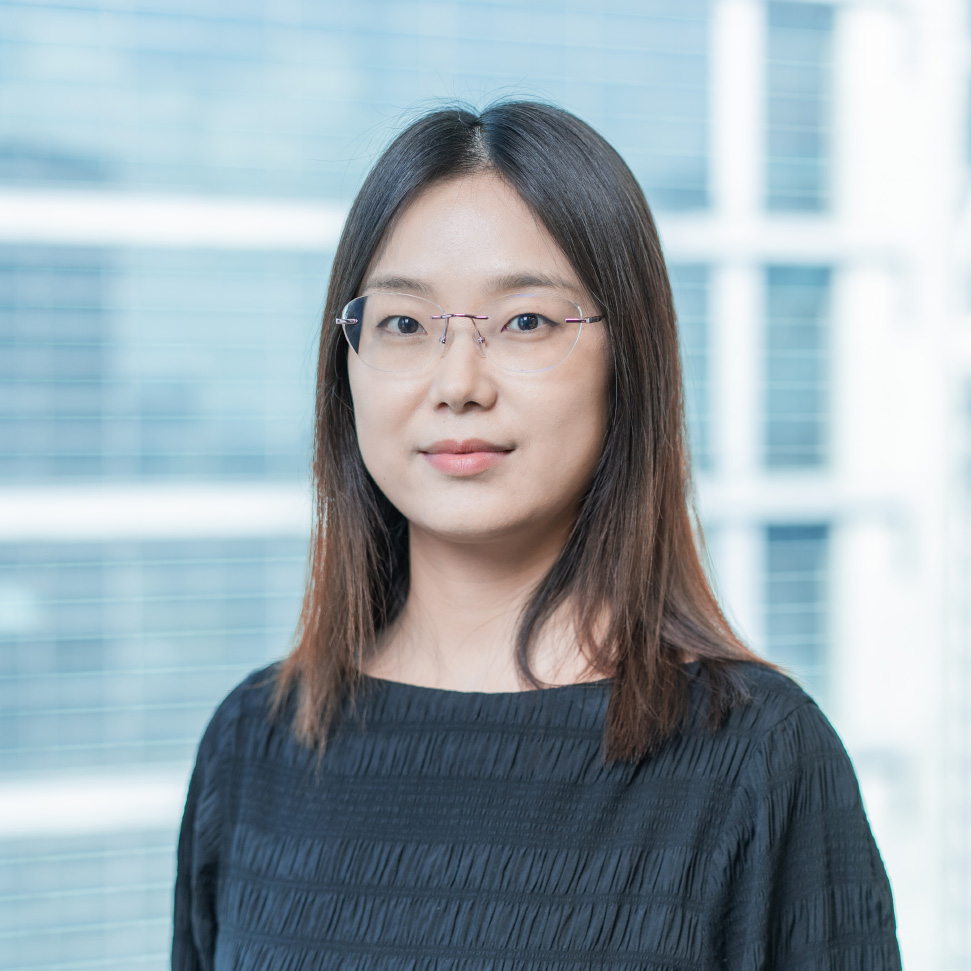 Headshot of a woman with shoulder-length dark hair and glasses, wearing a black top, standing in front of a softly blurred office background.