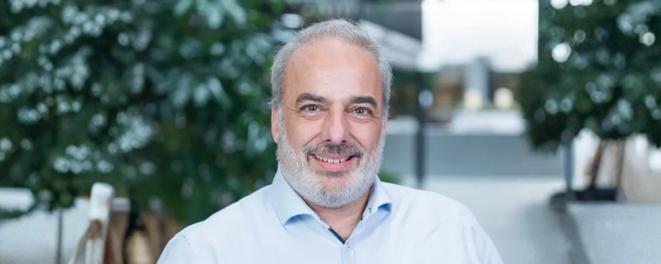 Headshot of Pascal Kocher, smiling, wearing a light blue button-down shirt, with a blurred greenery background.
