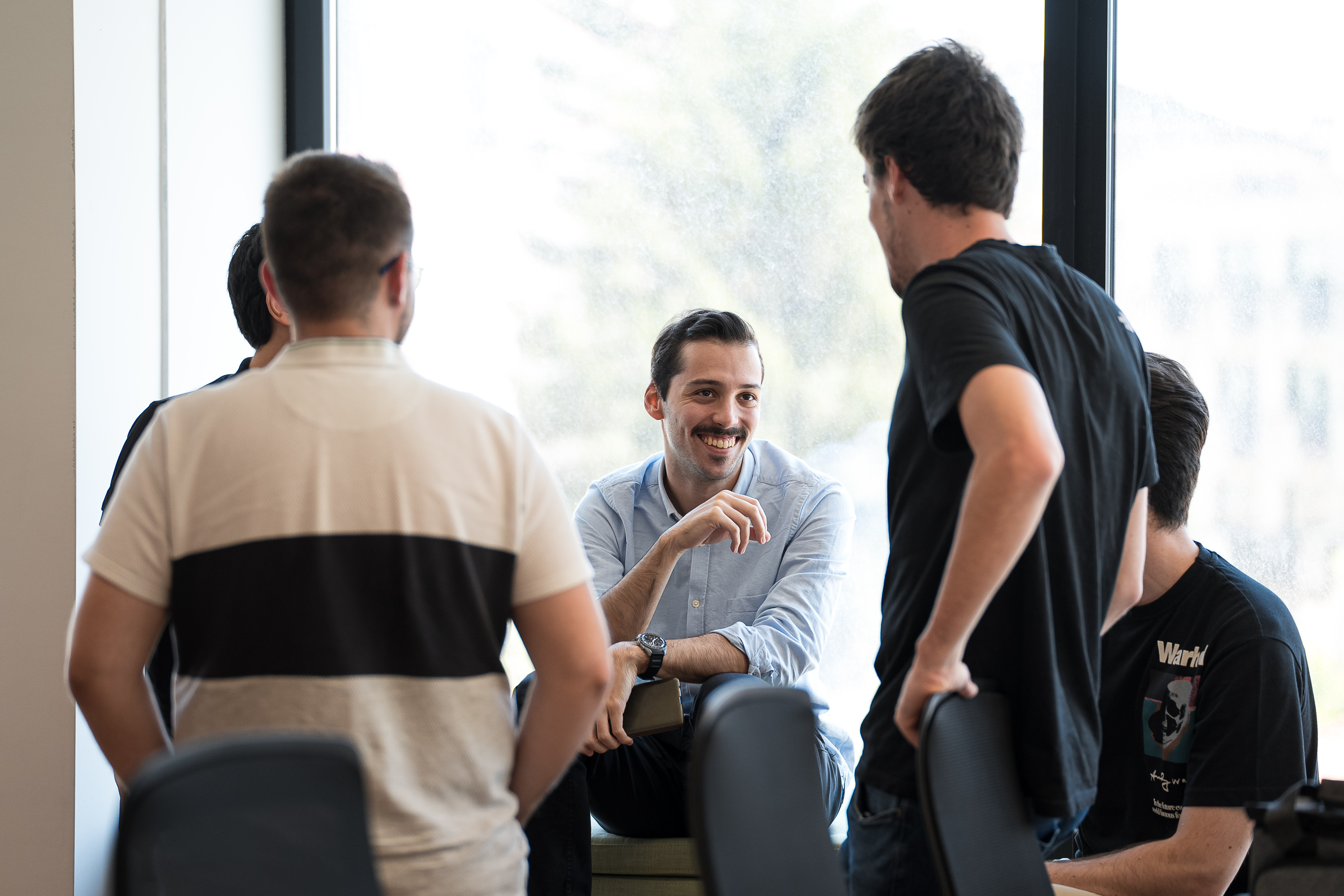 A smiling man sitting on an office window sill, around three male colleagues standing next to him