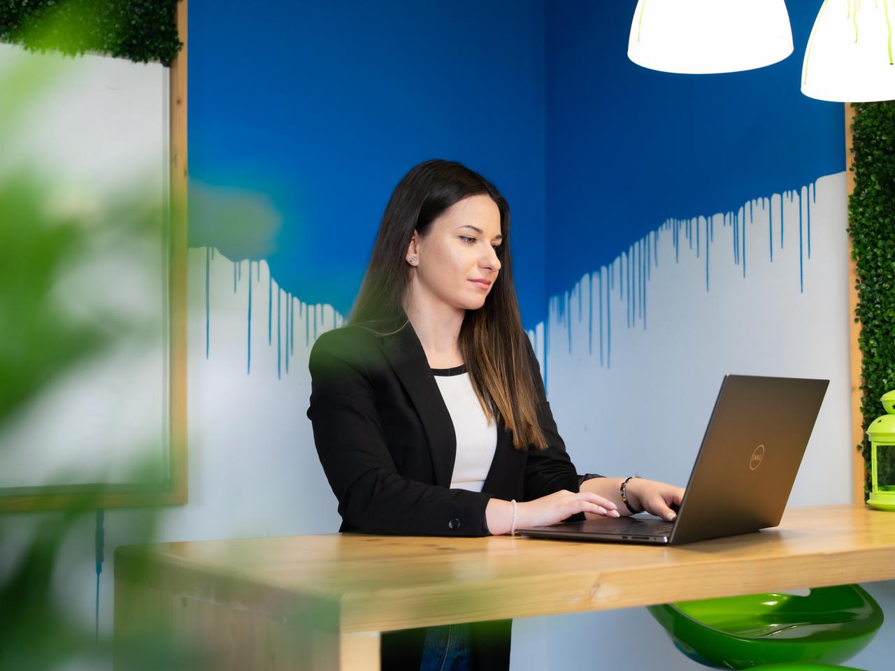 A smiling woman in a business suite standing in front of a laptop at a standing desk on a bright blue splash background in the office space