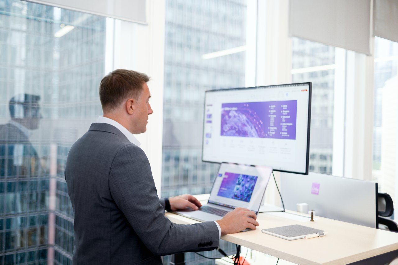 A colleague standing at the office desk in front of the laptop and a computer monitor, with the skyscraper buildings panorama in the background