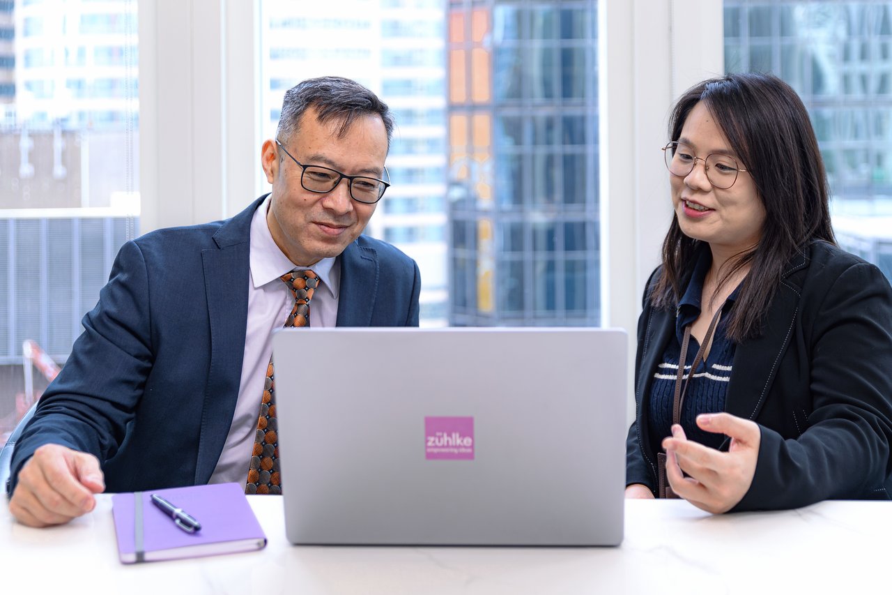 Two colleagues sitting at the office desk in front of the laptop, with the skyscraper buildings panorama in the background