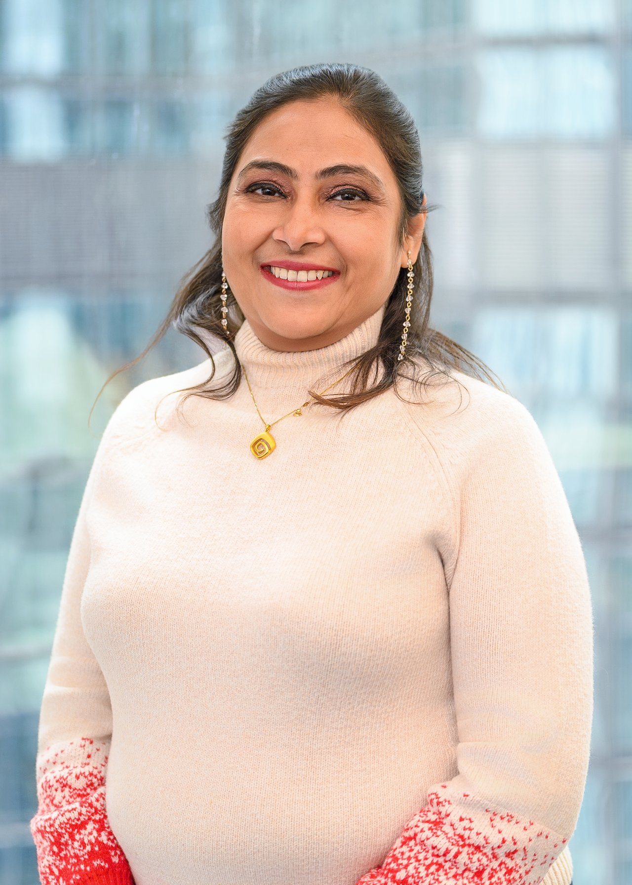 A smiling woman in a cream turtleneck jumper standing in the office space in front of the tall buildings skyline