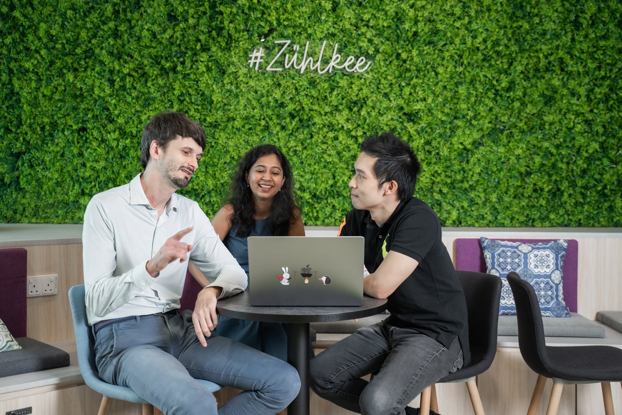 Three colleagues sit around a small table in a modern office lounge, discussing work while looking at a laptop. A green plant wall behind them displays the hashtag “#Zühlke,” creating a collaborative and friendly workplace atmosphere.