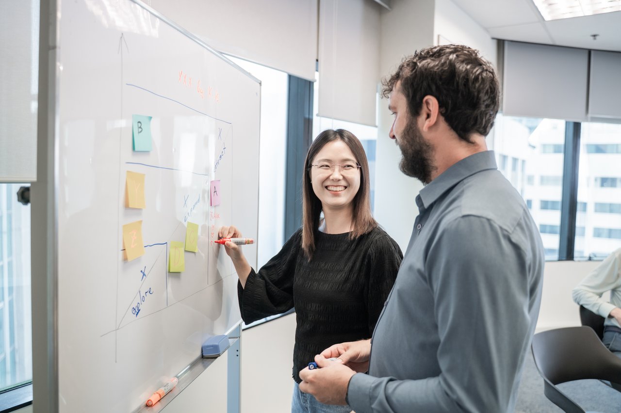 Two colleagues stand by a whiteboard in a bright office, smiling and discussing ideas. One person writes on the board with a marker while colorful sticky notes and sketches are visible, suggesting a collaborative planning or brainstorming session.