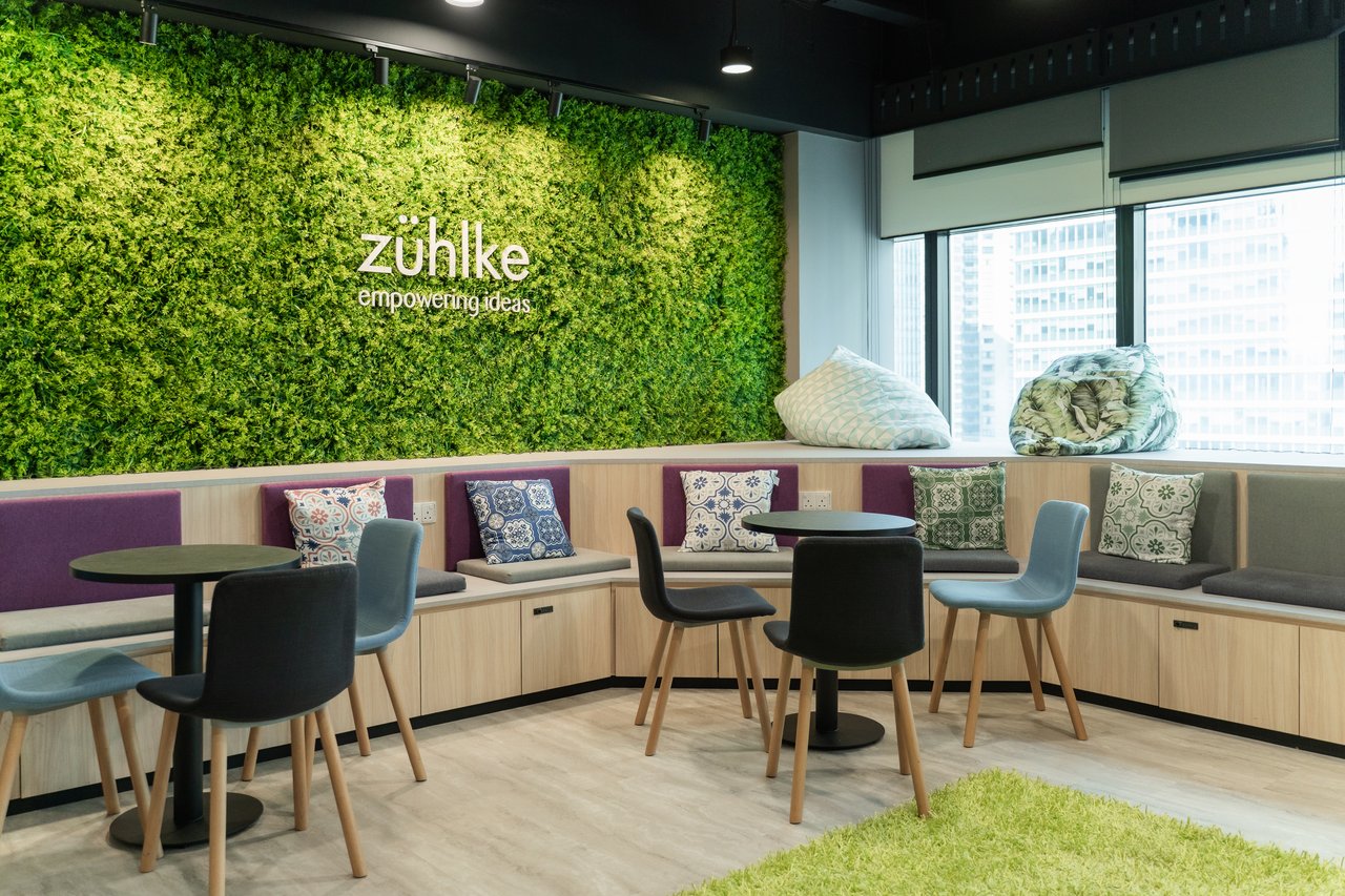 A modern office lounge with small round tables and chairs arranged around built-in seating. A large green plant wall displays the text “Zühlke – empowering ideas,” creating a welcoming and relaxed collaboration space with natural light from nearby windows.