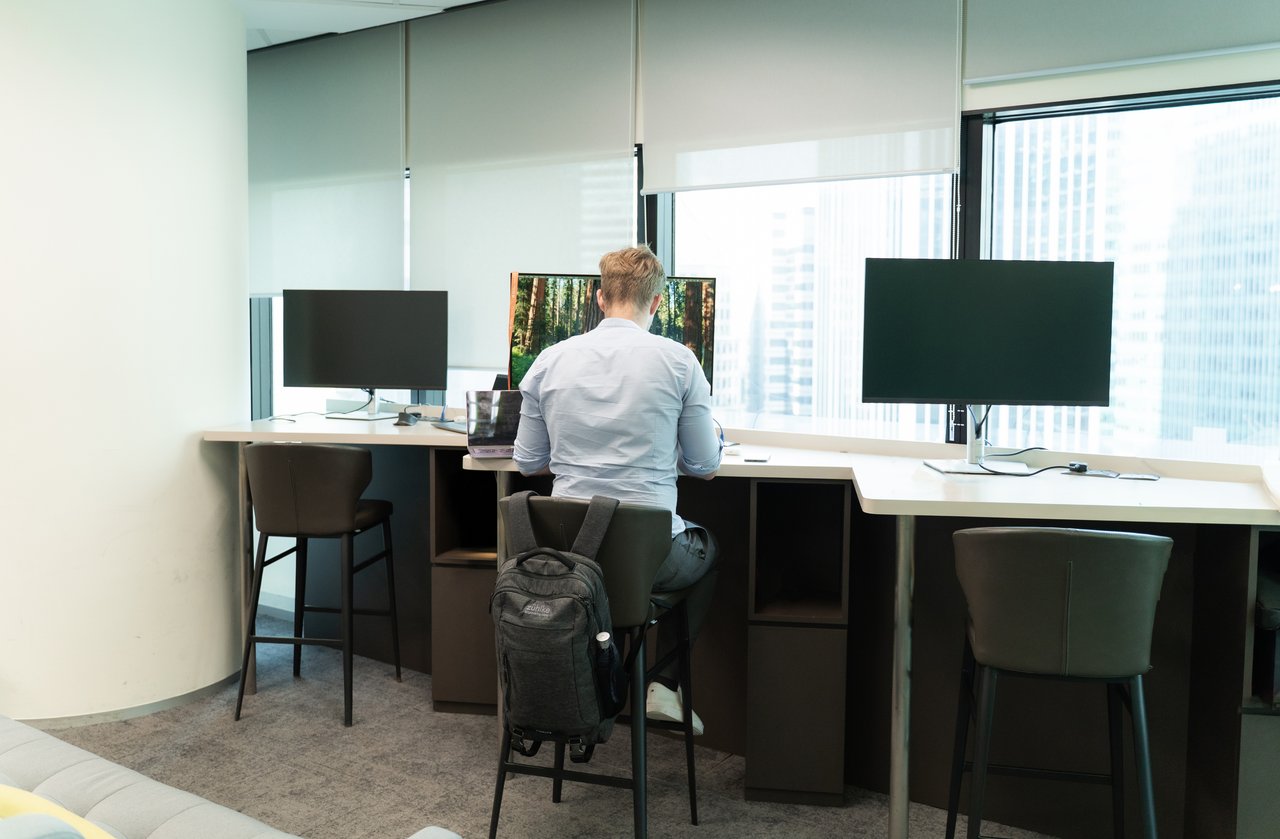 **Alt text:** A person sits on a high stool at a shared desk in a modern office, working on a laptop with multiple monitors nearby. Large windows let in natural light, and the workspace appears quiet and focused.