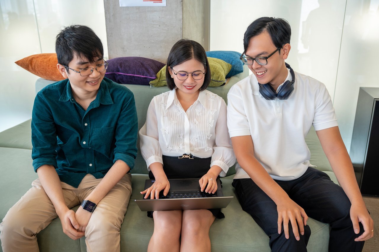 Three colleagues sit together on a sofa in a modern office lounge, smiling and collaborating while looking at a laptop. Colorful cushions and a relaxed setting suggest an informal team discussion or shared work session.