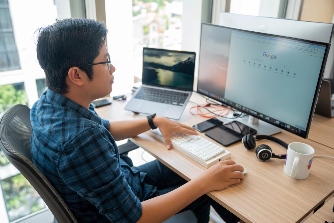 A tech professional siting at the office desk in front of a large monitor and a laptop