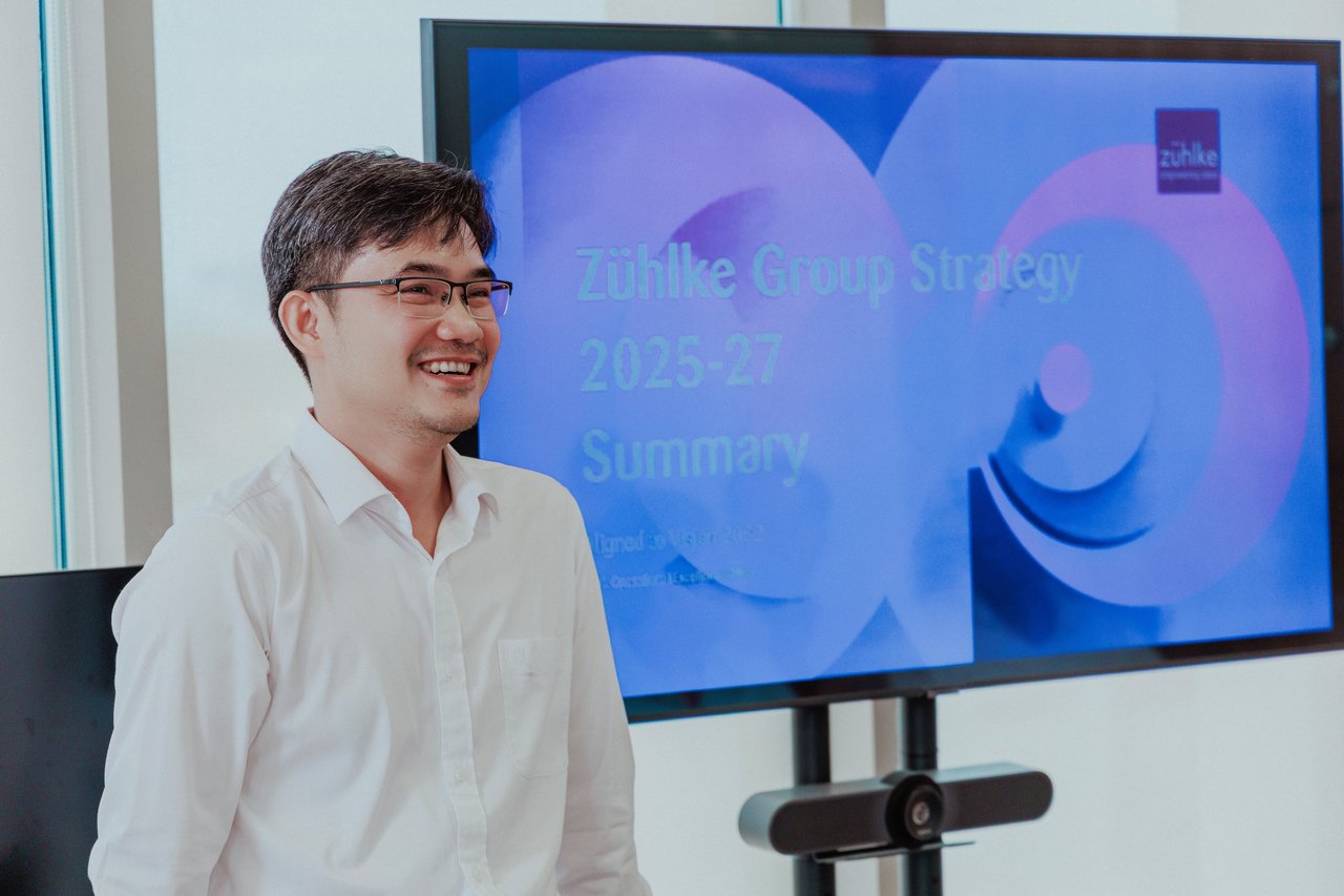A person stands smiling in front of a large presentation screen in a meeting room. The screen displays a slide titled “Zühlke Group Strategy 2025–27 Summary,” suggesting a professional presentation or briefing in progress.