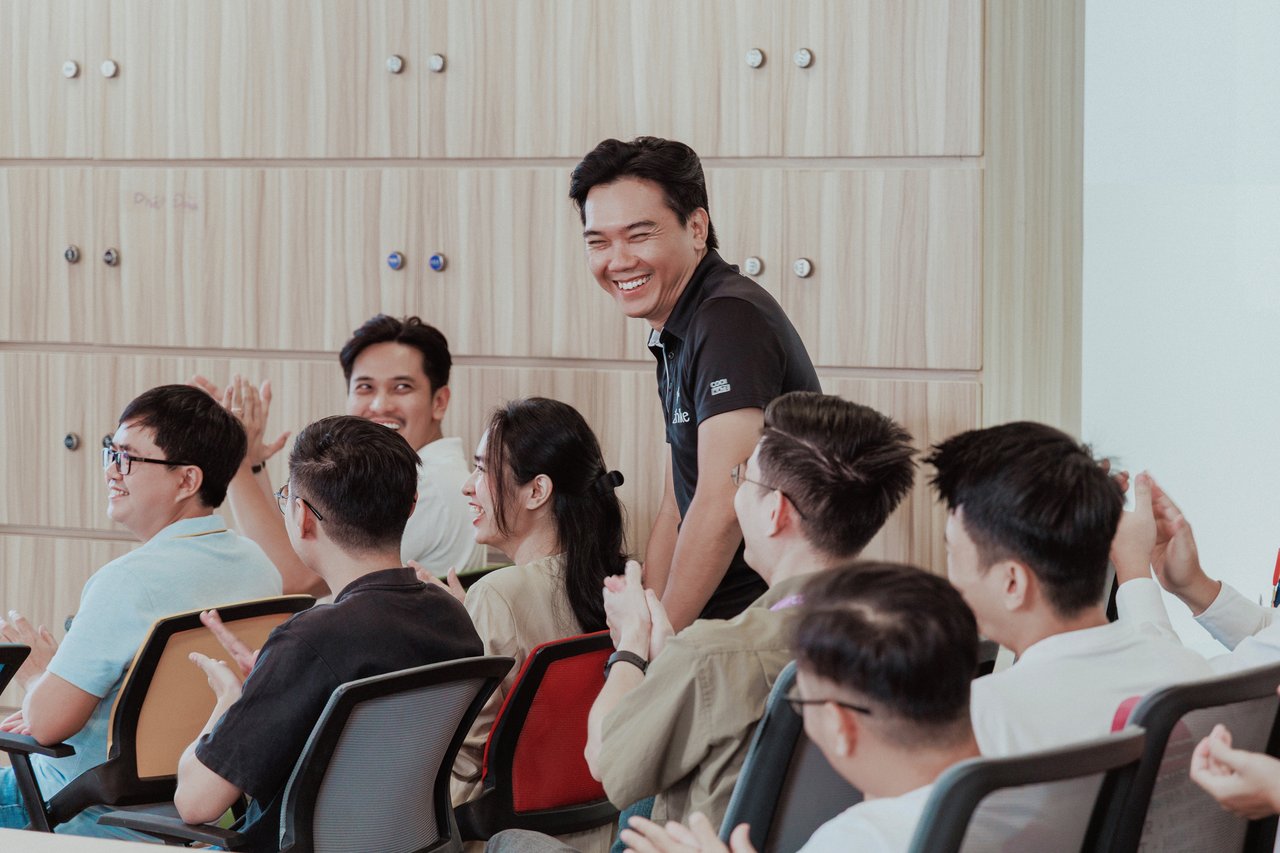 A group of colleagues sit together in a meeting room, smiling and applauding. One person stands among them, laughing, capturing a lively and positive moment of team engagement and celebration.