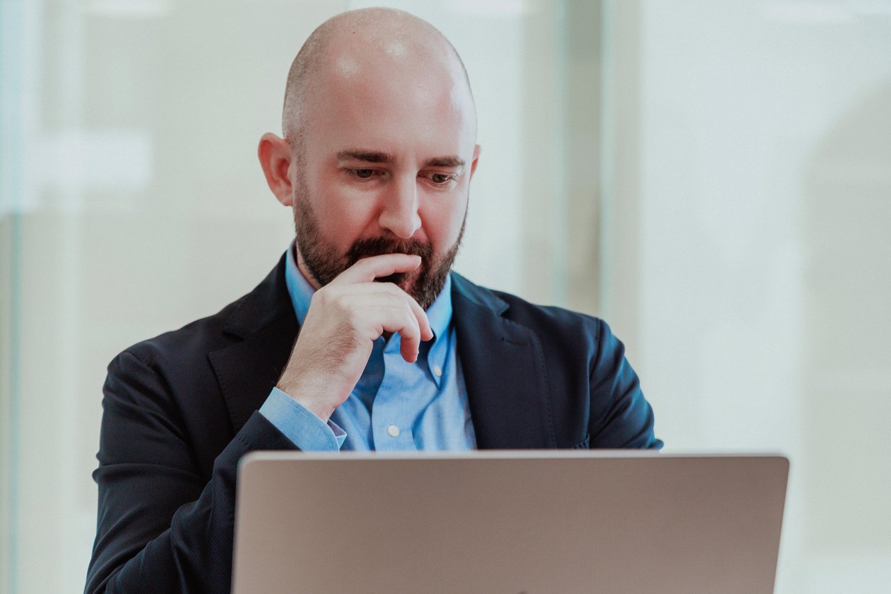 A person sits at a desk in a bright office, focused on a laptop screen. One hand rests thoughtfully near their face, conveying concentration and deep focus during work.