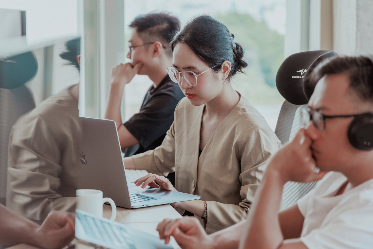 Several colleagues work together at a shared desk in a bright office. One person – a woman in the middle – focuses on a laptop while others sit nearby wearing headsets, creating a concentrated and collaborative working atmosphere.