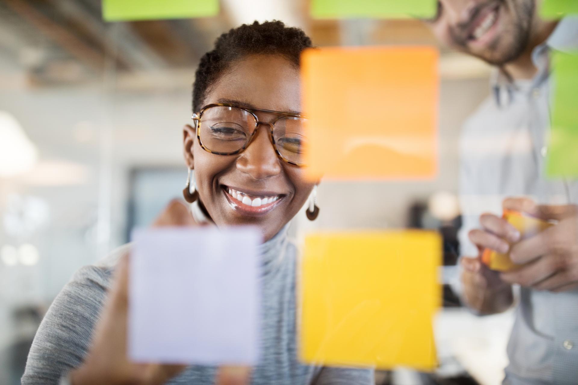 Smiling businesswoman brainstorming during an office workshop, writing ideas on colourful sticky notes on a glass wall.