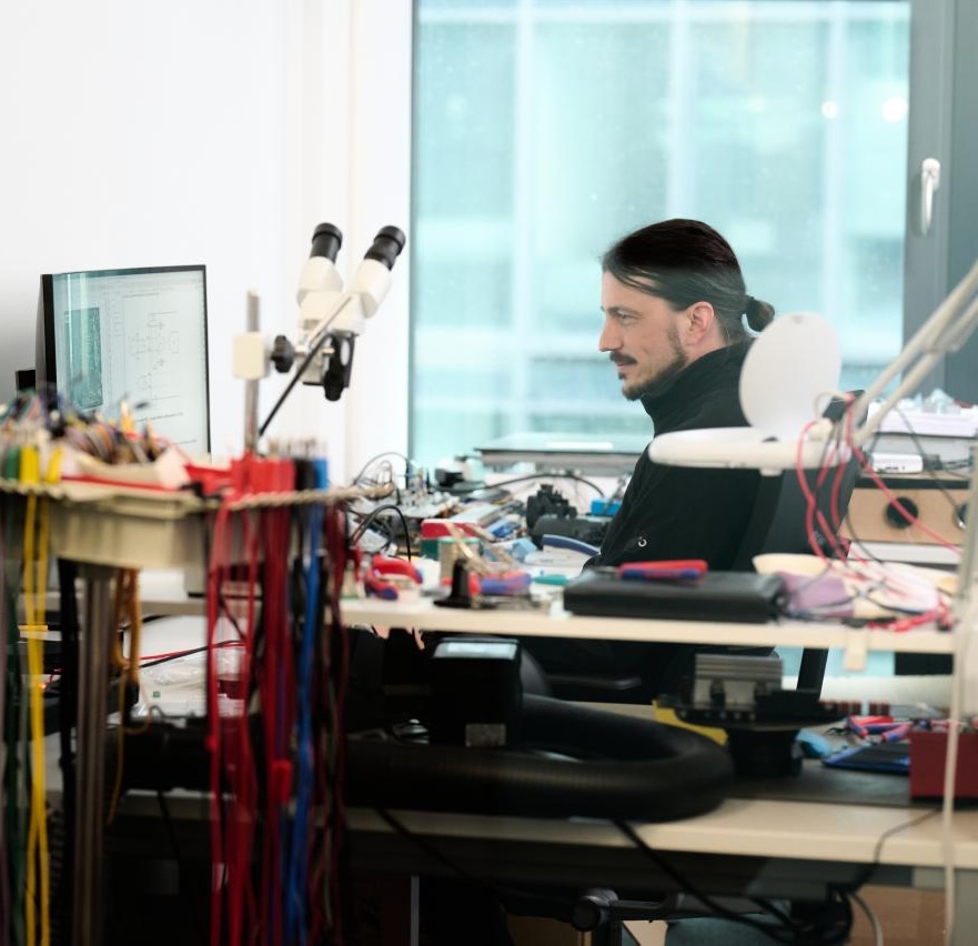 A hardware engineer sits at a workbench, working at a monitor surrounded by electronic components, wiring harnesses, and lab equipment including a binocular microscope, illustrating the hands-on engineering work behind hardware development.