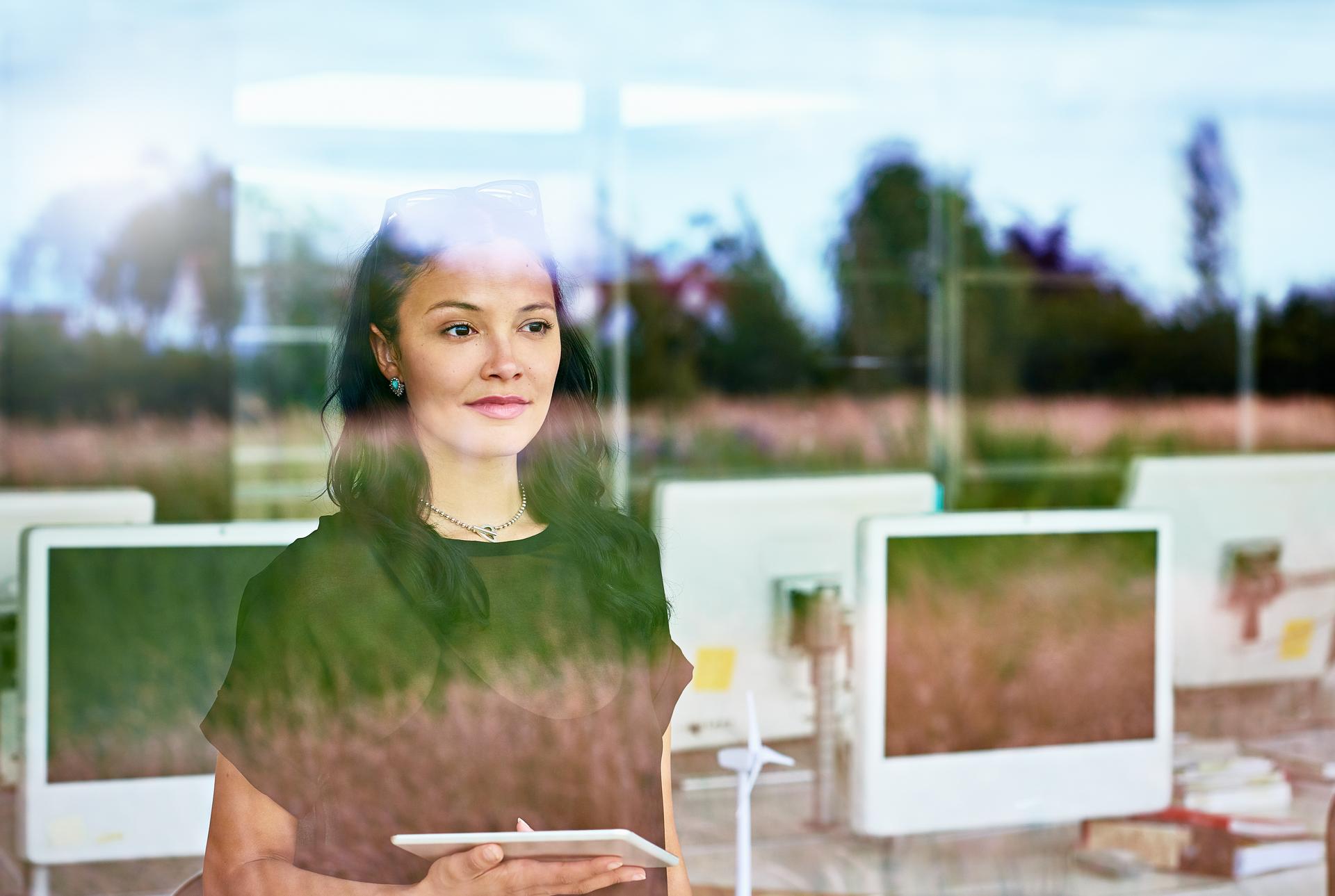 Businesswoman holding a tablet, looking out through office window with reflections, suggesting future-focused strategy and planning.
