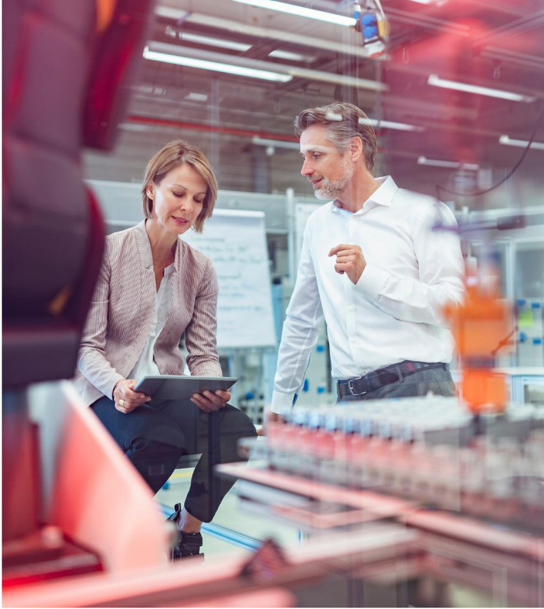 Colleagues reviewing data on a tablet on a factory floor, with production equipment and machinery in the background