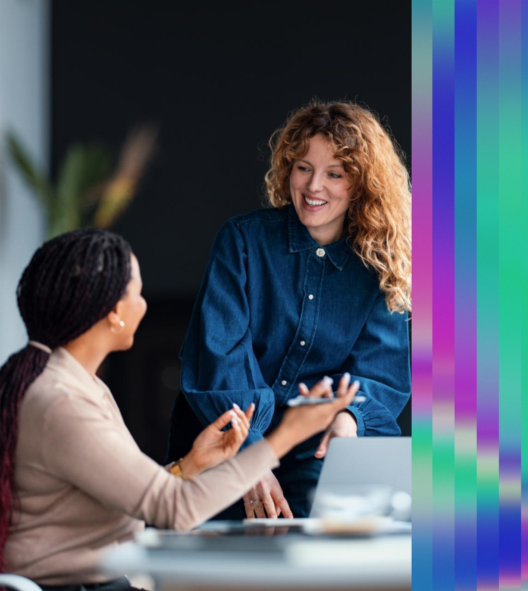 Two colleagues in a modern office setting engaged in a friendly discussion; a woman with curly hair leans forward smiling while another woman gestures as she speaks, with a laptop on the table and a soft, blurred background.