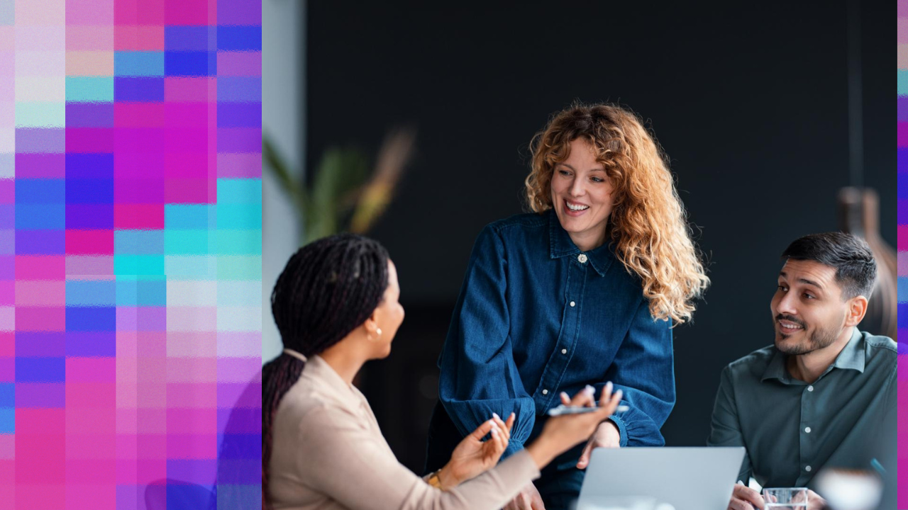 Three colleagues collaborating around a table in an office; a woman with curly hair smiles while listening to a seated colleague who is gesturing, while a third colleague looks on, with a laptop and drinks visible, conveying a positive team interaction.