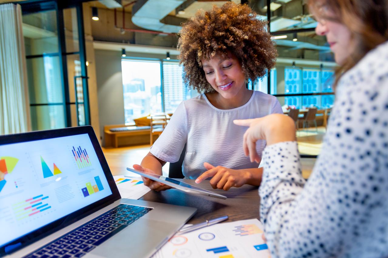 Two colleagues in an office reviewing finance charts and graphs on a laptop and tablet, one pointing to a chart as they discuss the data together.