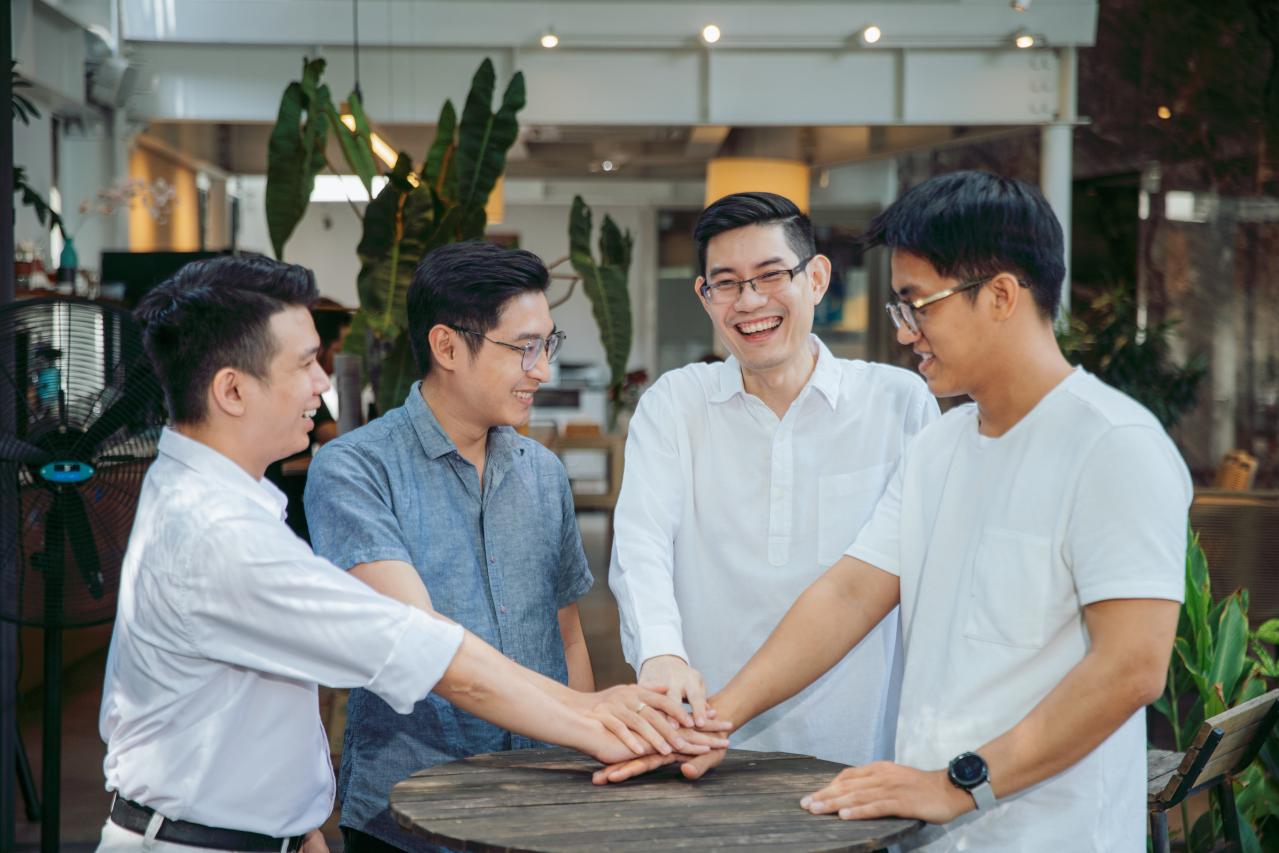 Four colleagues smiling and stacking their hands together around a table in the office.
